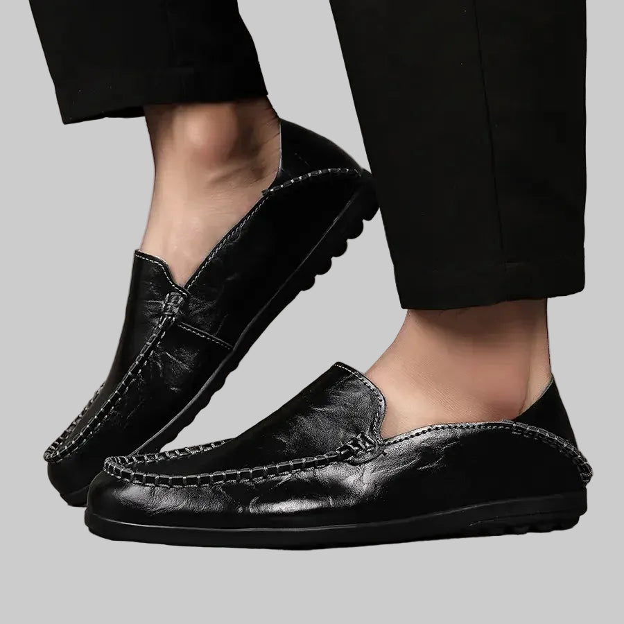 Black leather loafers worn with black pants on a wooden floor.
