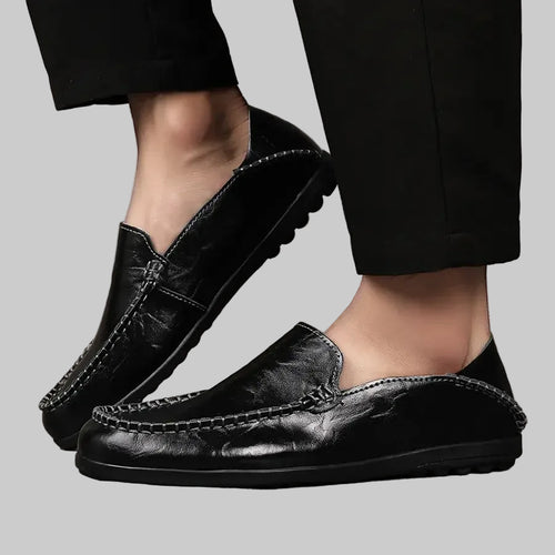 Black leather loafers worn with black pants on a wooden floor.